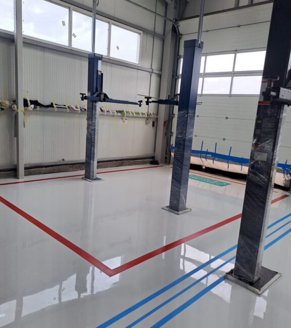 Pardoseală epoxidică lucioasă alb-gri cu marcaje roșii și albastre într-un service auto.